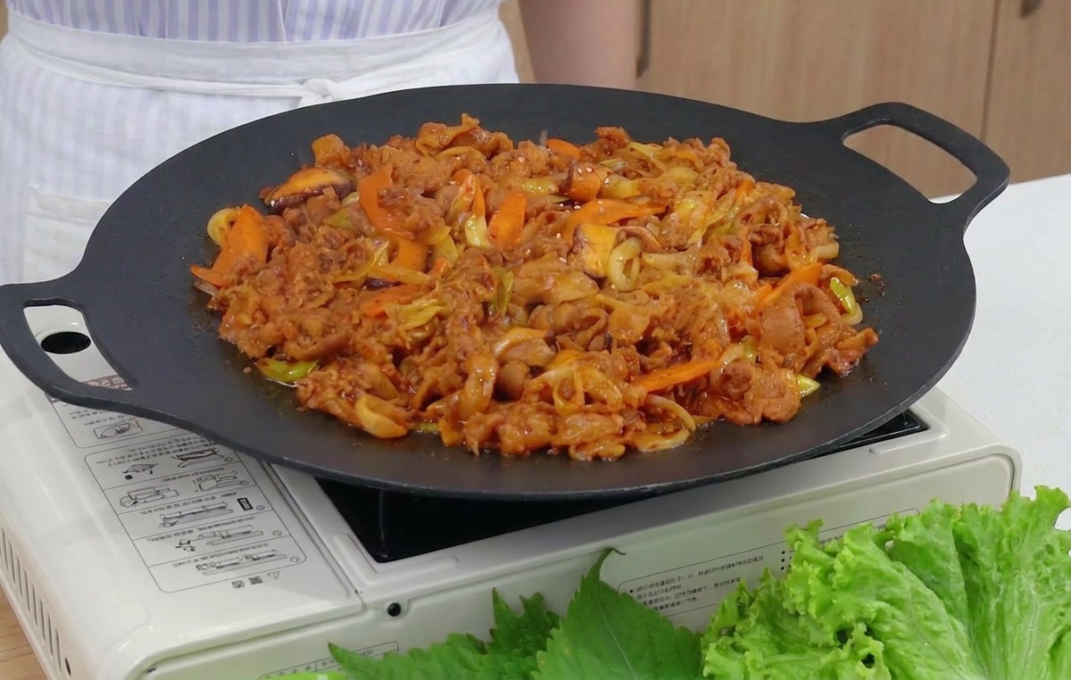 A close-up shot of toasted sesame seeds being sprinkled over a steaming pan of red, spicy stir-fried pork and vegetables.