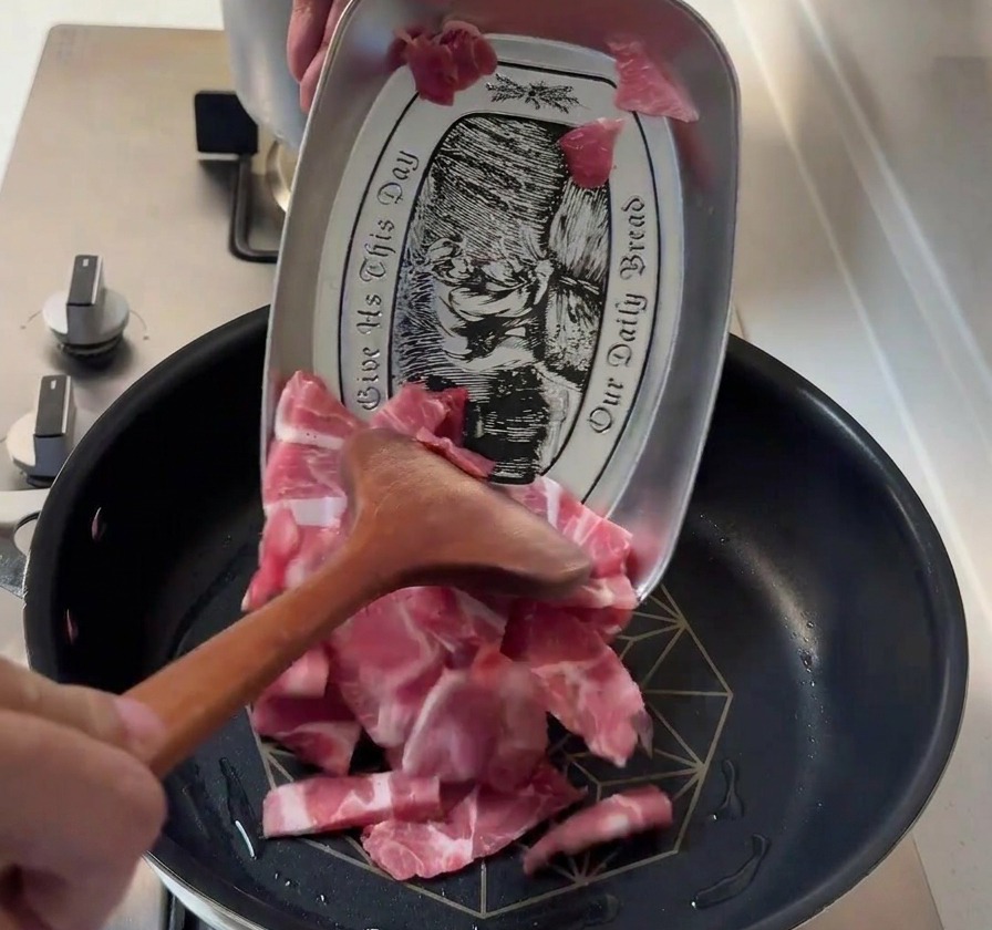 Thin slices of raw pork shoulder being added to a hot pan from a patterned metal tray using a wooden spatula.