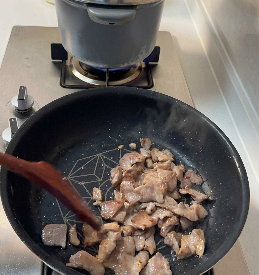Sizzling pork shoulder pieces with golden-brown edges being stirred in a dark frying pan.