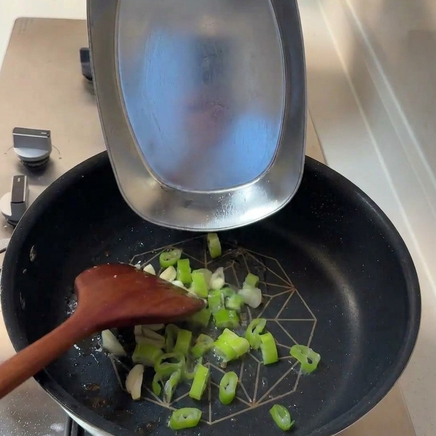 Chopped scallions and sliced garlic cloves being added to a pan with residual cooking oil.