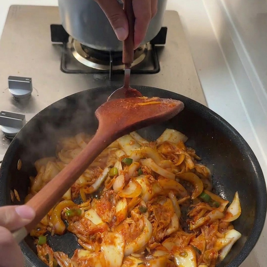 A spoonful of thick red gochujang paste being added to the pan of stir-frying ingredients.