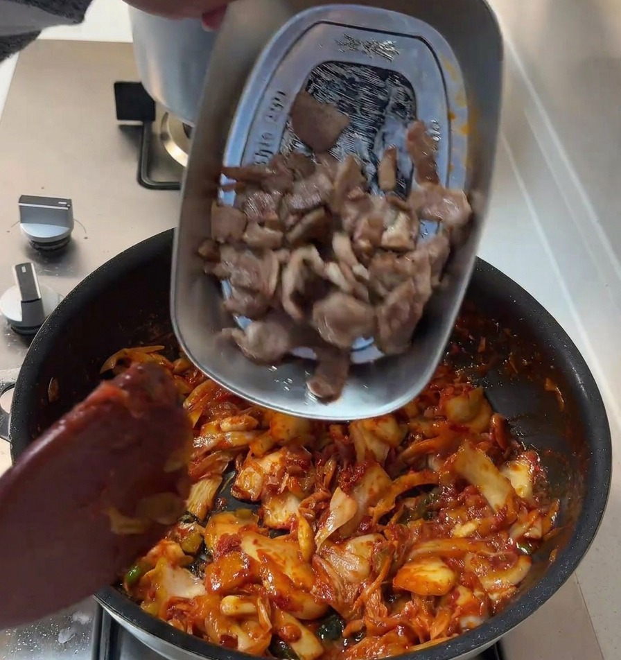 Seared pork slices being poured from a metal tray into a pan containing a red, spicy kimchi and onion mixture.