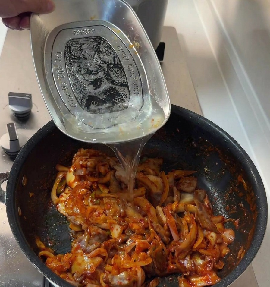 Water being poured from a metal tray into the pan of spicy pork and kimchi to finish the sauce.