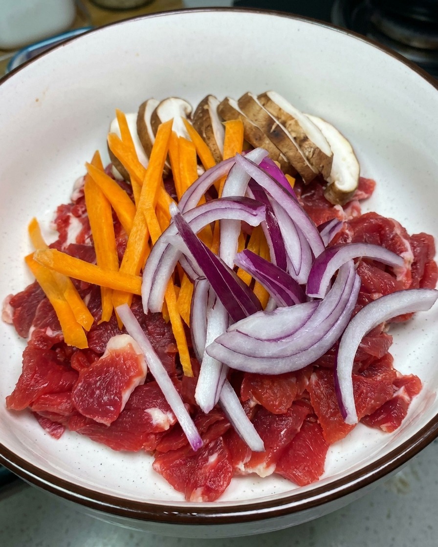 A white bowl filled with raw red beef slices topped with fresh sliced shiitake mushrooms, orange carrots, and red onions.