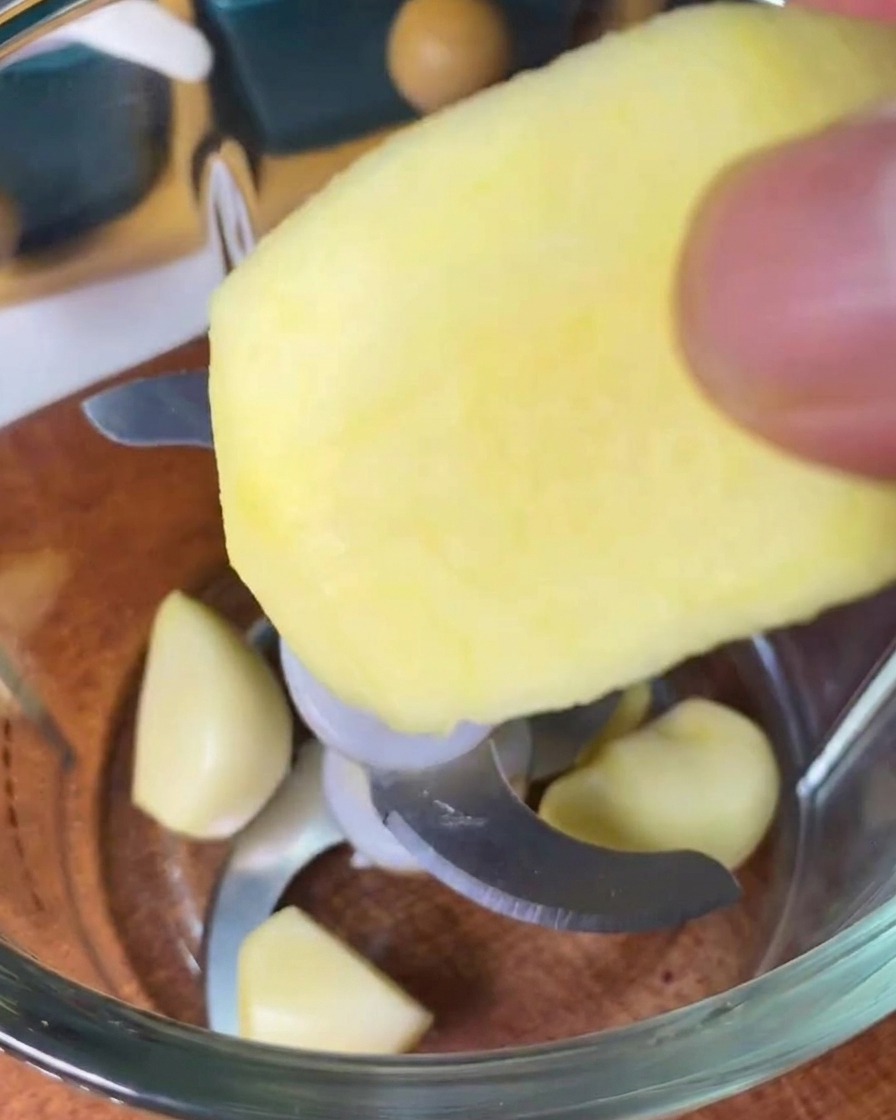 A person dropping a slice of peeled apple into a blender container that already contains several whole garlic cloves.