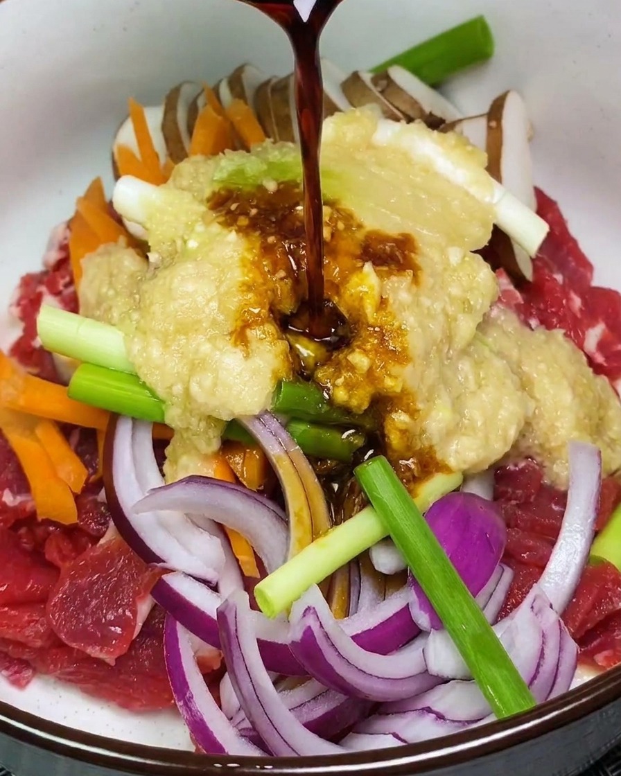 Dark marinade liquid being poured onto a pile of raw beef and vegetables topped with a light-colored fruit puree in a white bowl.
