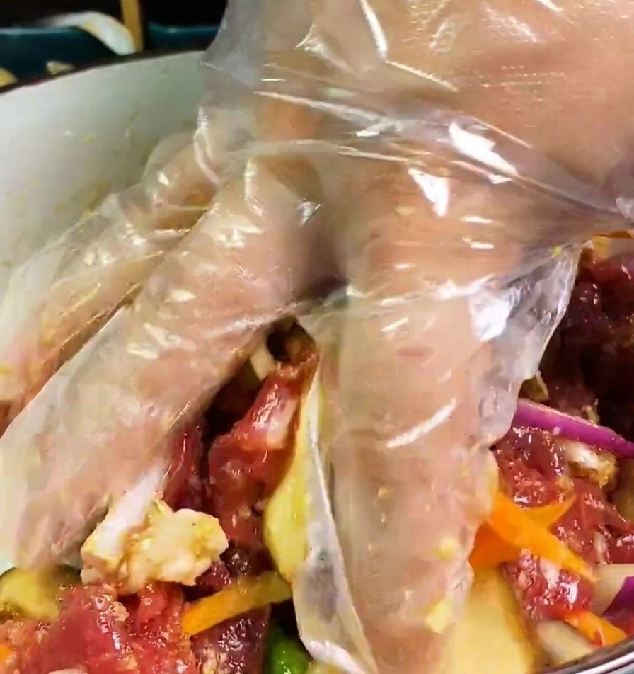 A hand in a clear plastic glove mixing together raw beef, sliced onions, carrots, and mushrooms in a bowl of dark marinade.