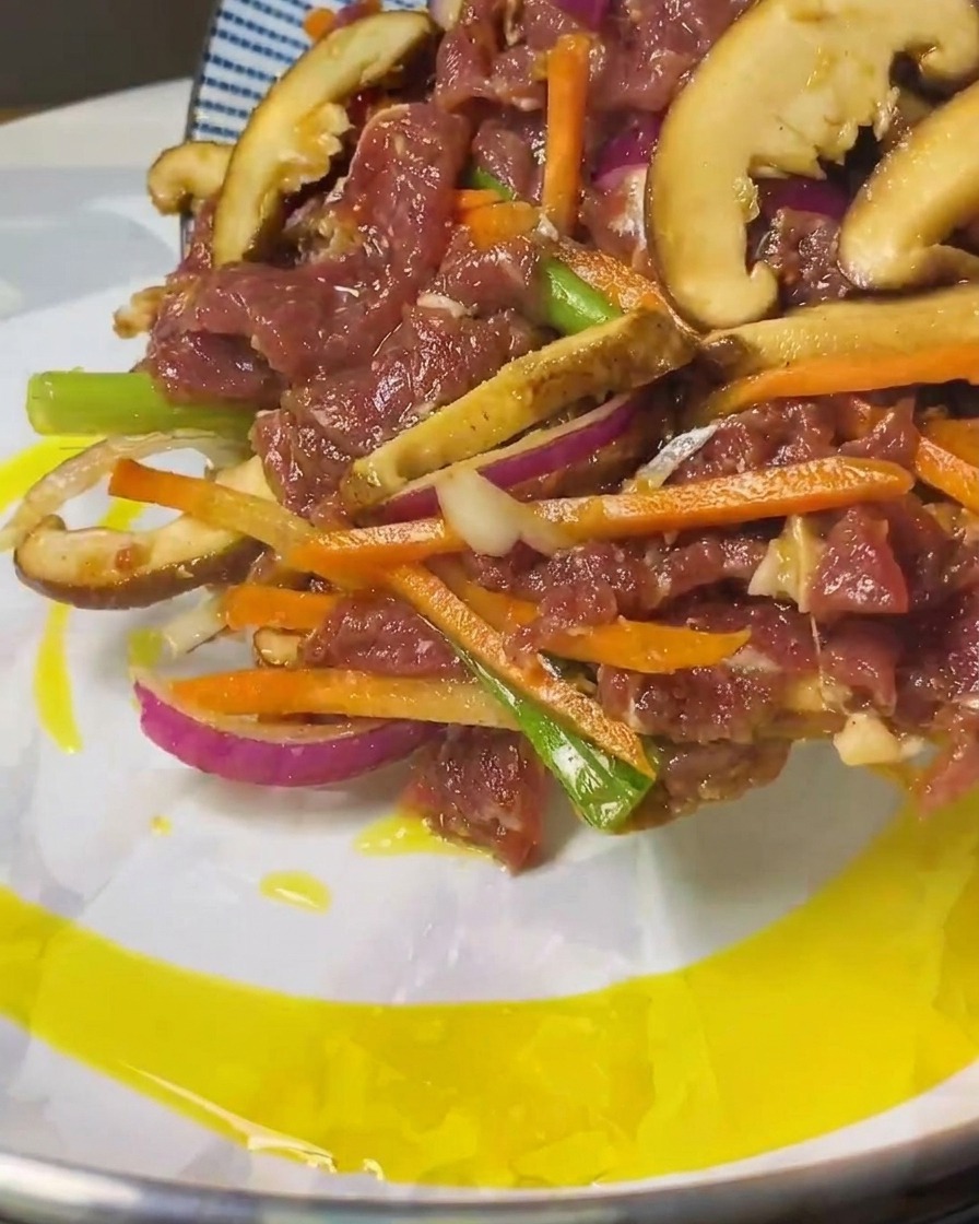 Marinated raw beef and vegetable strips being slid from a blue-patterned plate into a pan with heated oil.