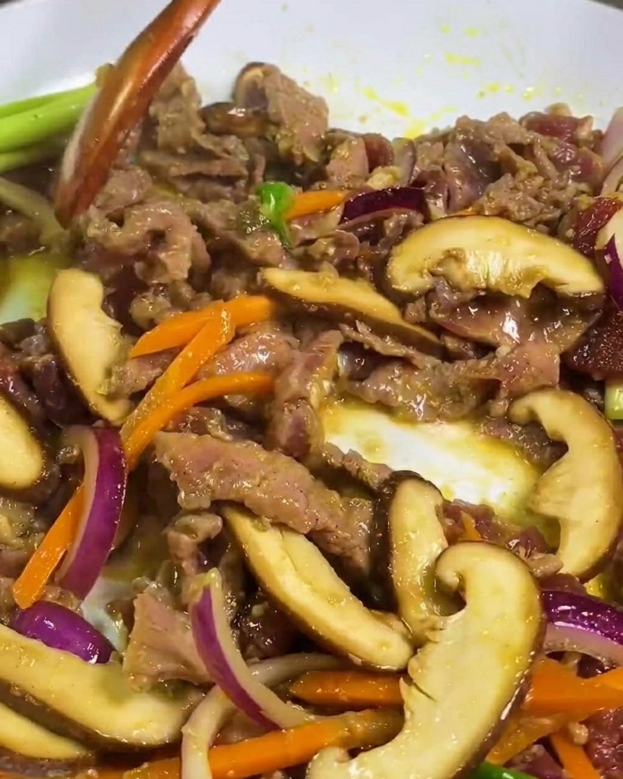 Thinly sliced beef, shiitake mushrooms, and carrots being stir-fried in a white pan with a wooden spatula.