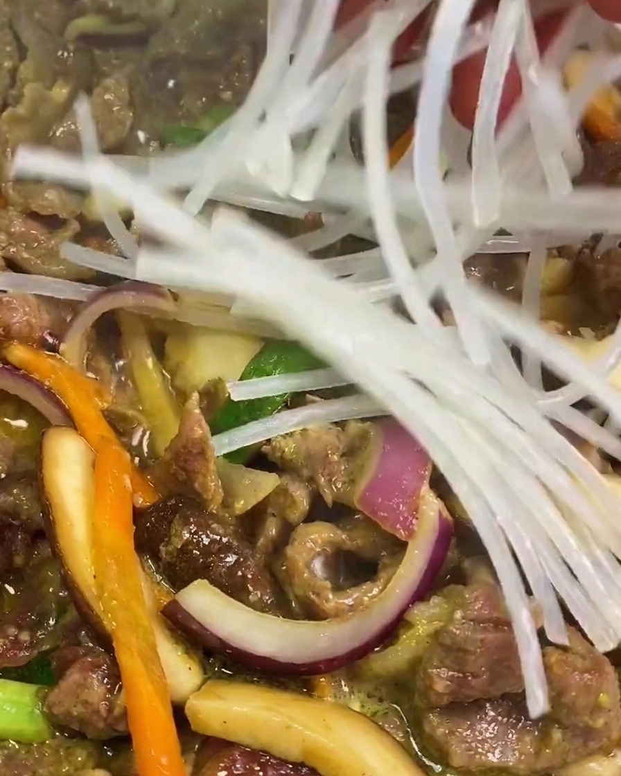 A hand dropping a bunch of clear, soaked sweet potato starch noodles into a pan of simmering beef and vegetables.