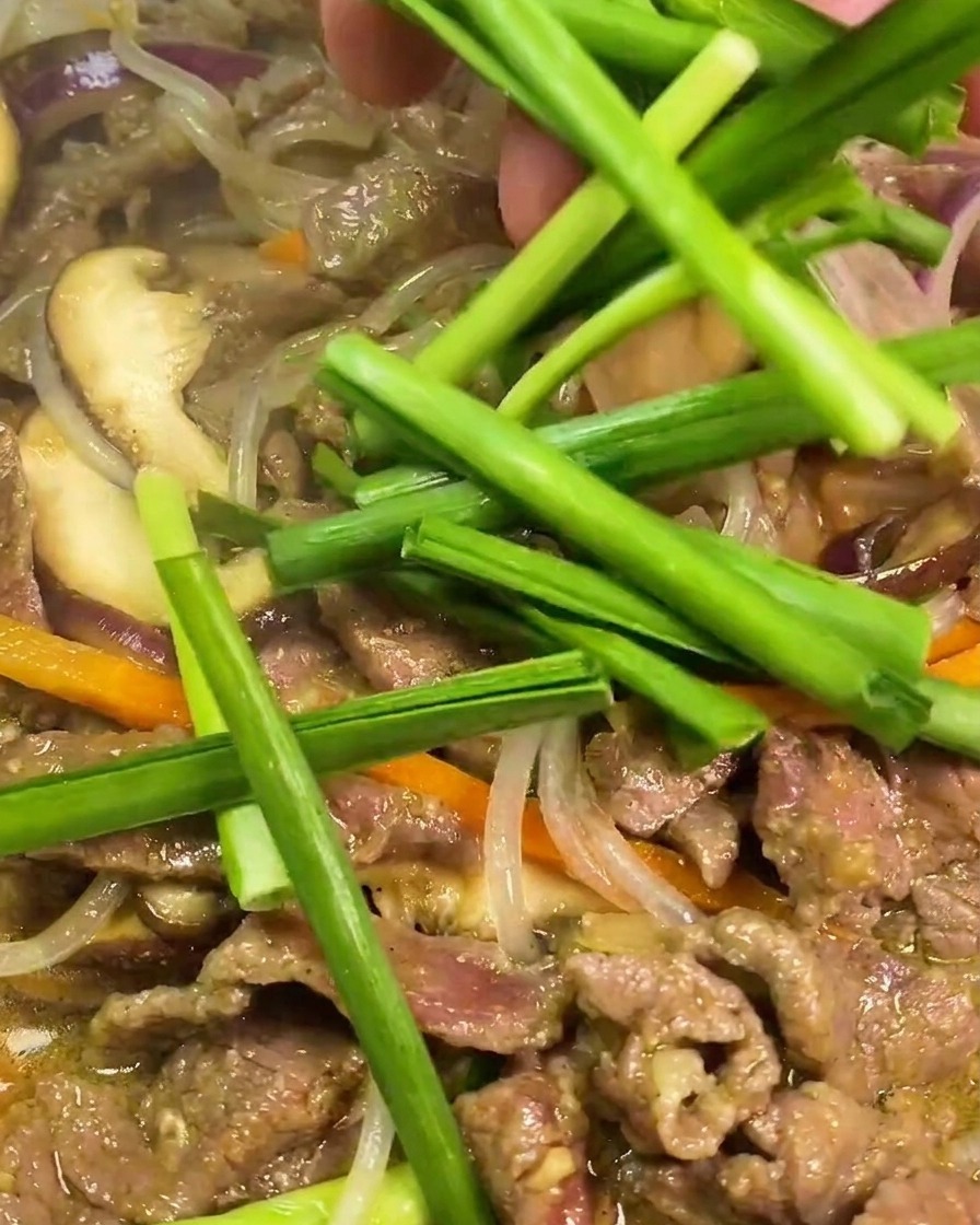 Fresh green Chinese chive segments being added by hand into a hot pan containing stir-fried beef, translucent glass noodles, sliced carrots, and purple onions.