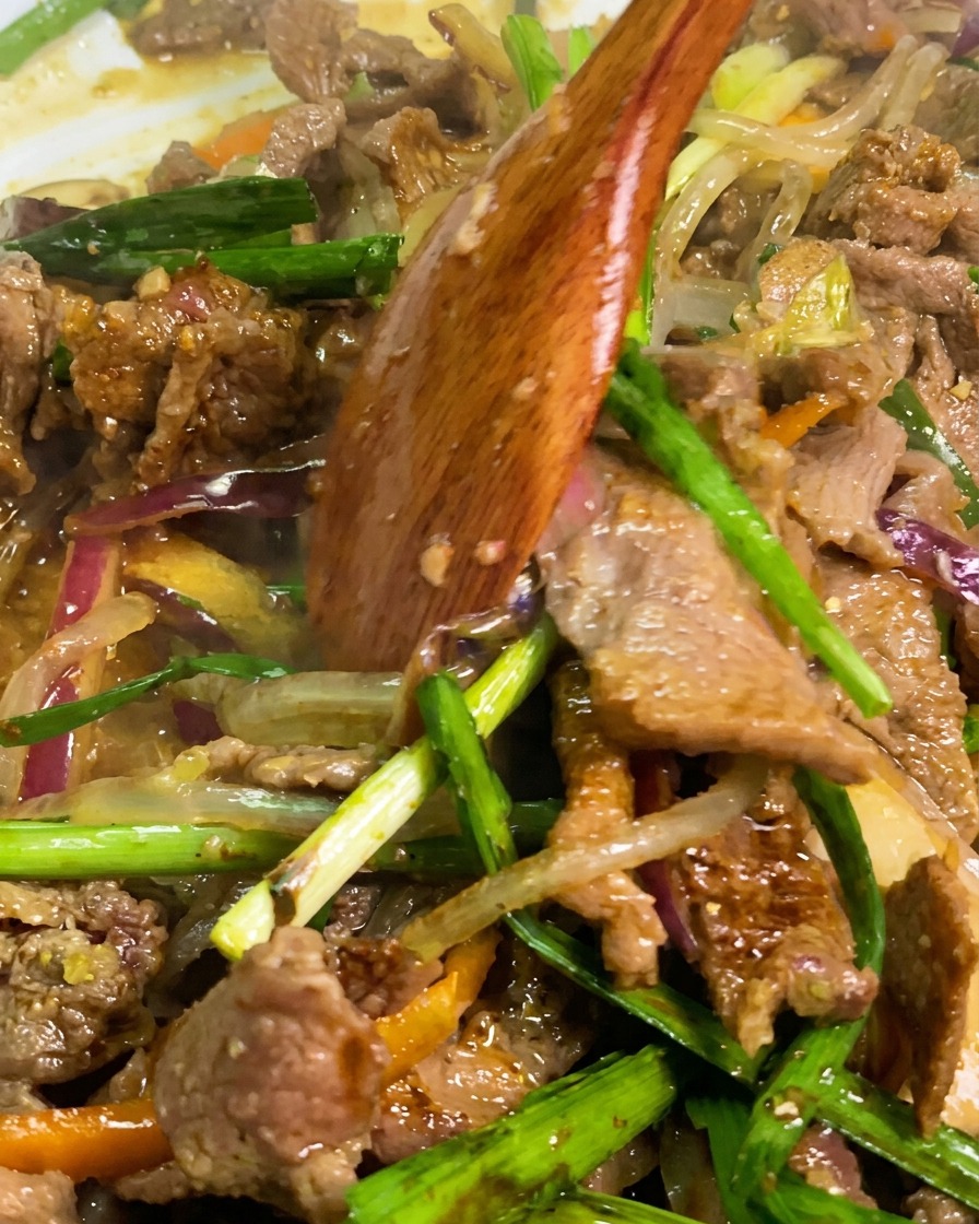 A wooden spatula mixing together beef, translucent starch noodles, and green chive segments in a pan.