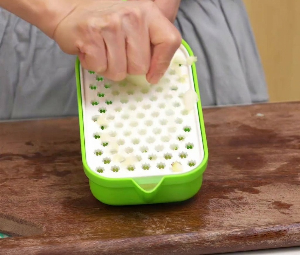 A person using a green plastic grater to turn a peeled apple into fruit pulp on a wooden cutting board.