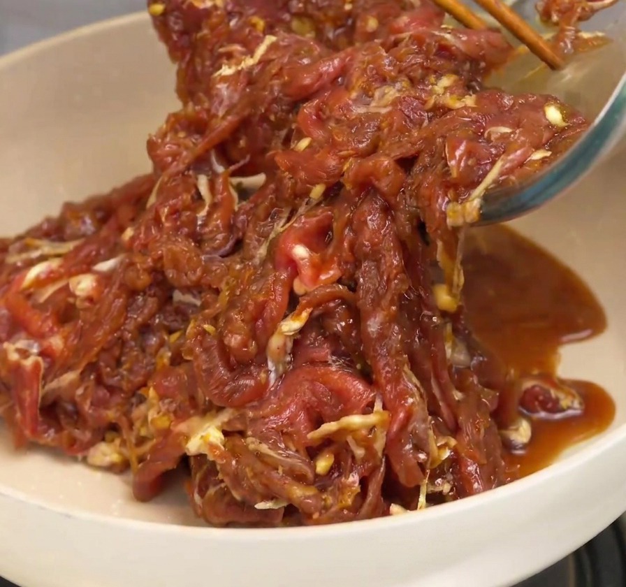 Marinated beef strips being poured into a white ceramic frying pan and stirred with wooden chopsticks.
