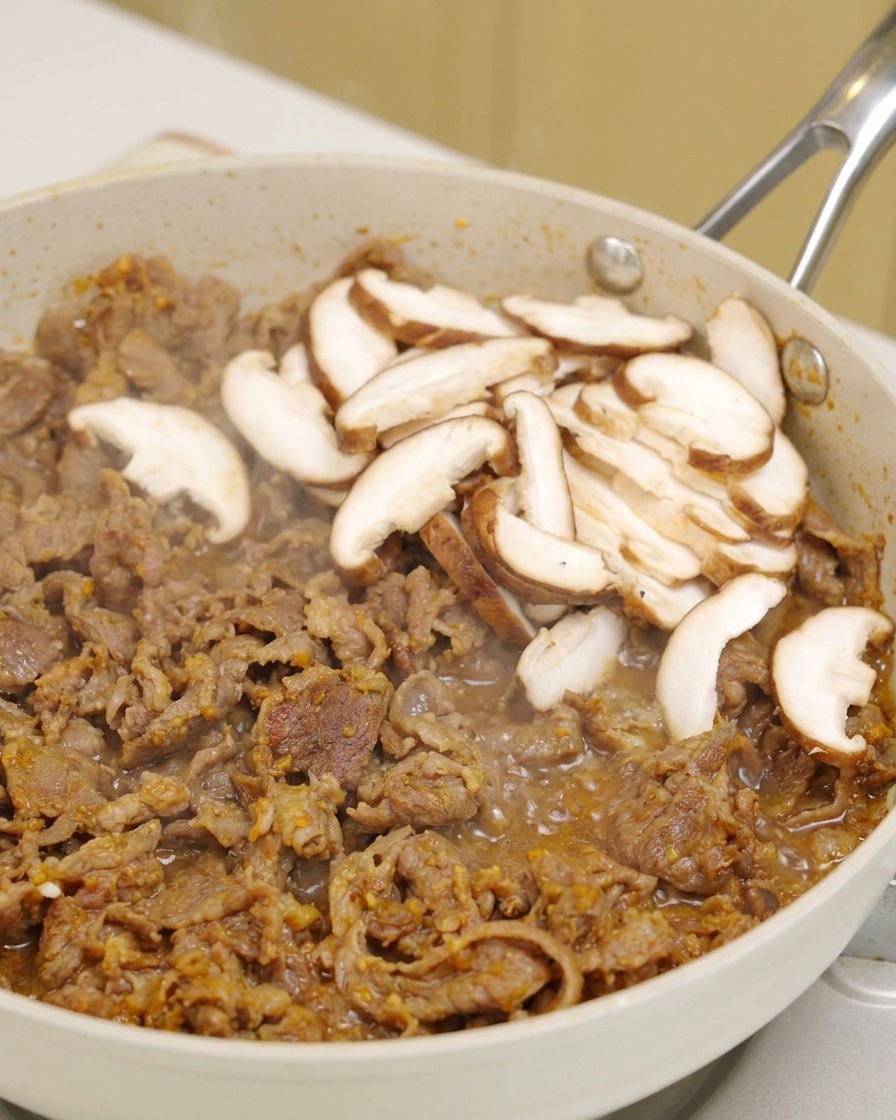 Slices of fresh shiitake mushrooms being placed on top of sizzling beef in a white pan.