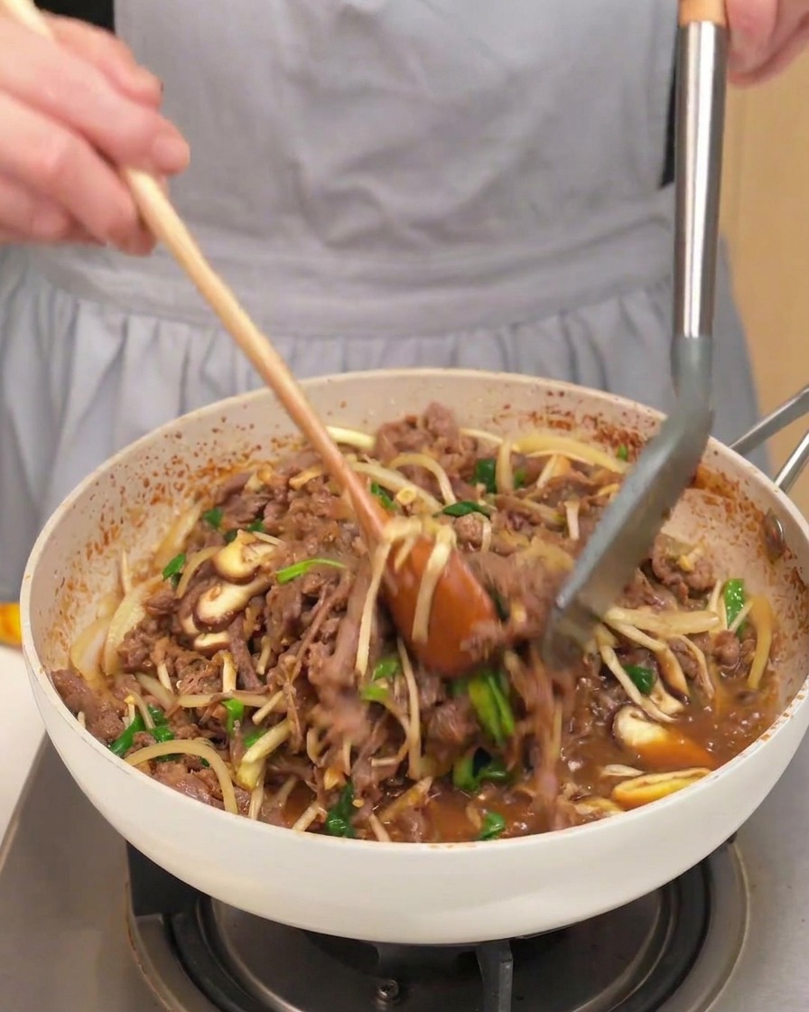 A pan full of cooked beef, mushrooms, onions, and green scallions being tossed with a wooden spoon and tongs.