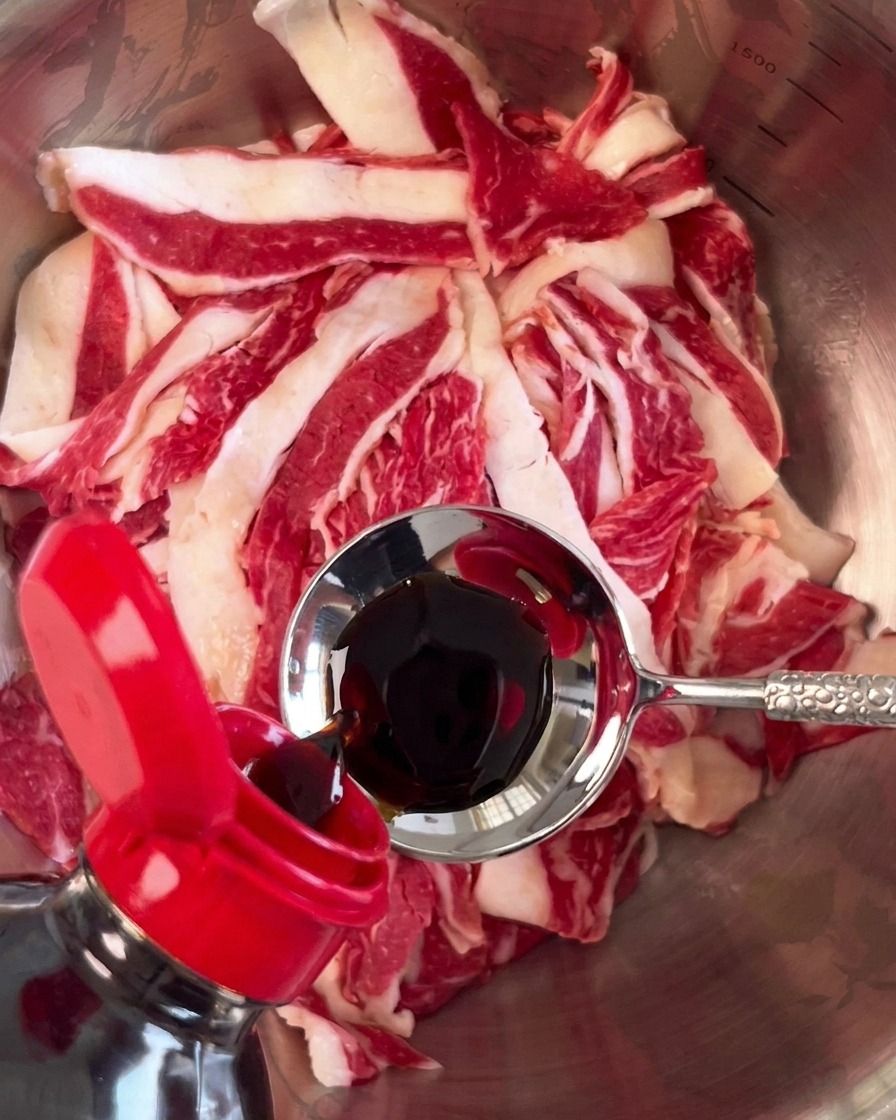 Dark soy sauce being poured from a metal spoon onto a pile of raw sliced beef in a silver bowl.