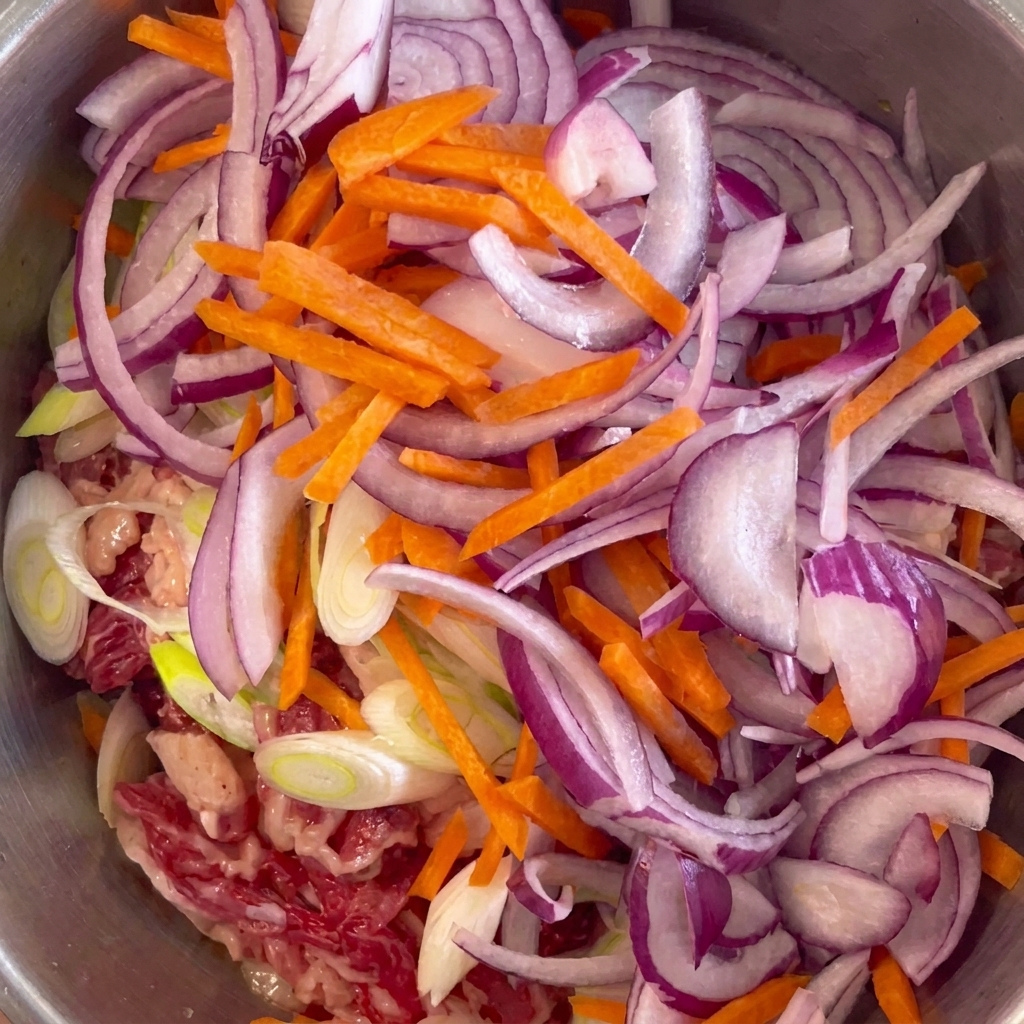 Sliced red onions and bright orange carrot matchsticks arranged on top of marinated beef in a stainless steel bowl.