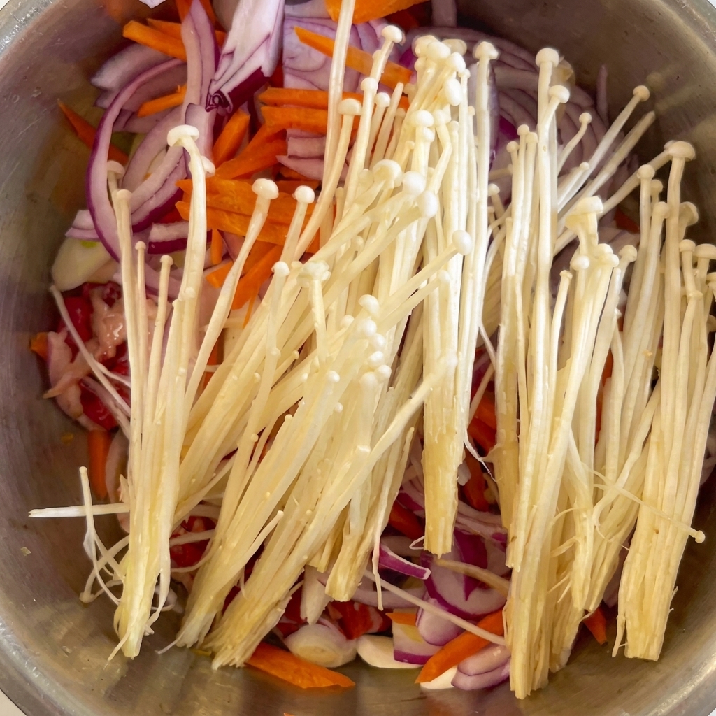 Fresh white enoki mushrooms and minced garlic cloves placed over the beef, onions, and carrots.