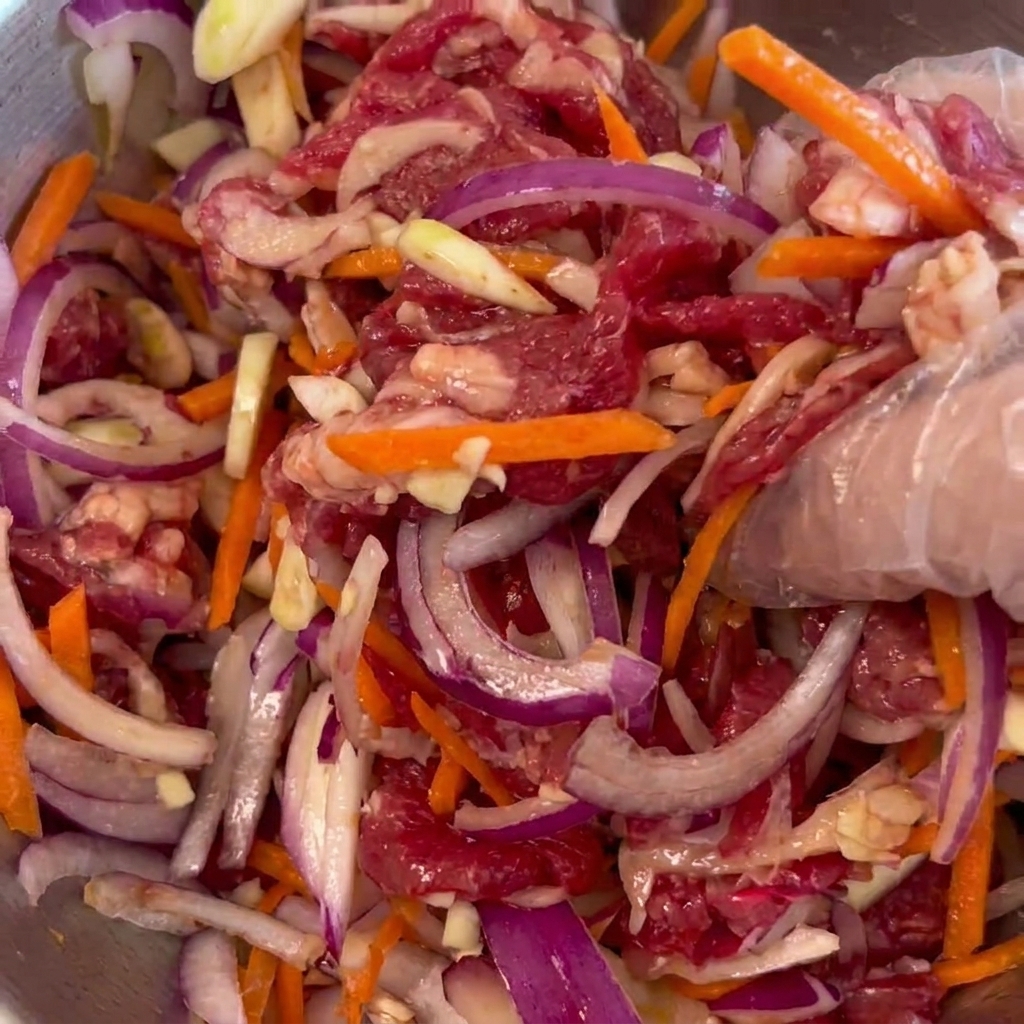 A hand wearing a clear plastic glove mixing together raw beef, purple onions, and carrot sticks in a metal bowl.