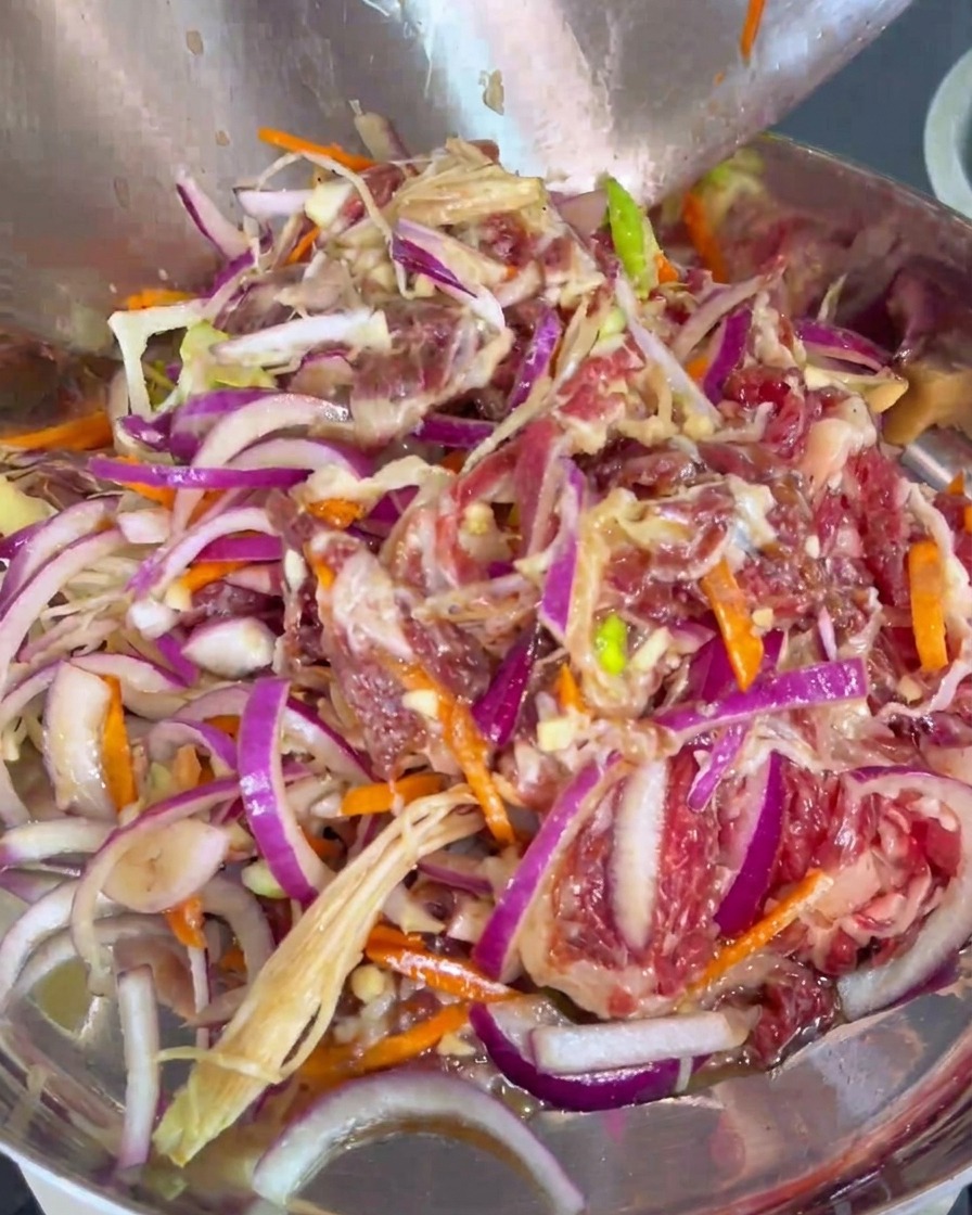Raw marinated beef slices thoroughly mixed with red onions and carrots being transferred into a metal cooking pan.