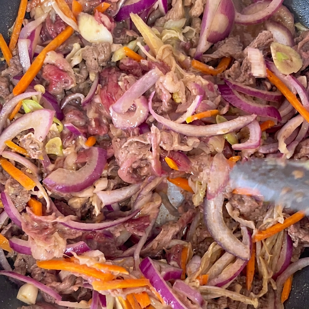 Marinated beef strips and assorted sliced vegetables cooking in a hot frying pan.