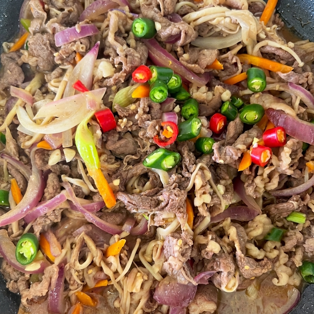 Brightly colored sliced red and green chilies resting on top of cooked beef bulgogi in a pan.