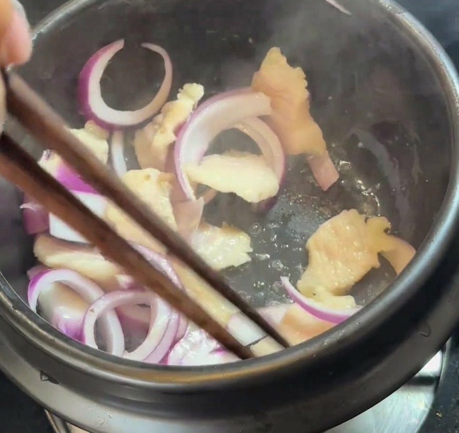 Fresh red onion slices being mixed into the rendered fat and pork belly pieces in a pot.