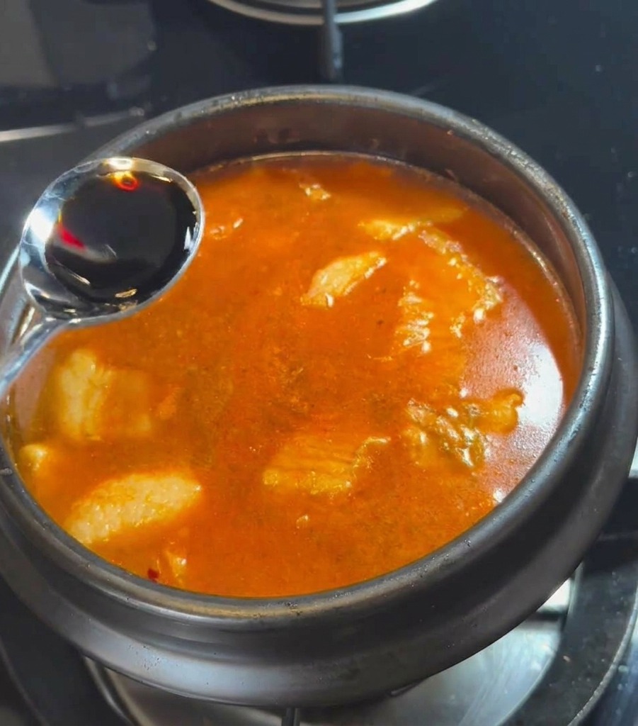 A silver spoon pouring dark soy sauce into the bubbling red soup broth.