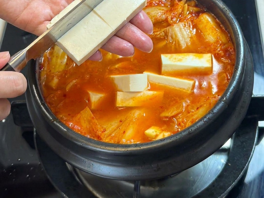 A person using a knife to slice a block of white tofu directly over a bubbling pot of spicy kimchi stew.