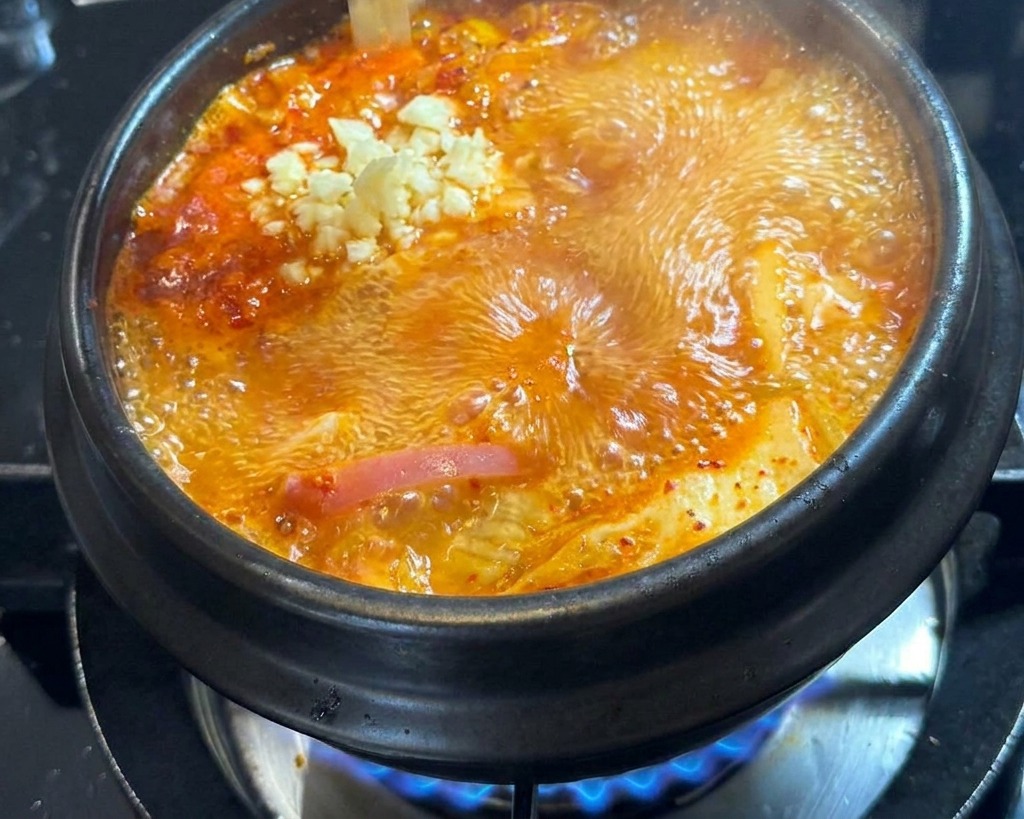 Fresh minced garlic being dropped from a hand into the center of a boiling pot of red soup.