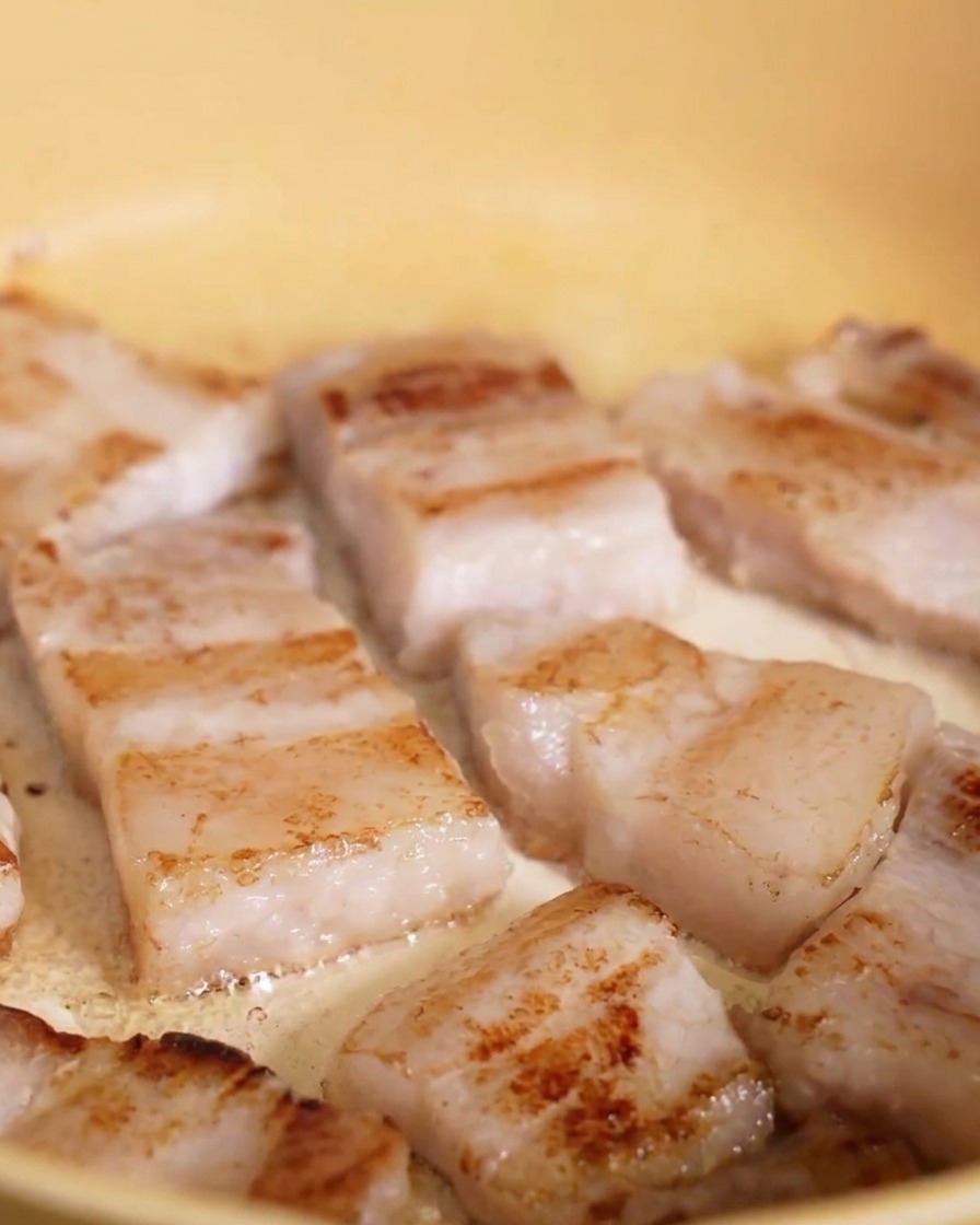 Slices of pork belly frying in a yellow pan, with the undersides showing a distinct golden-brown crust.