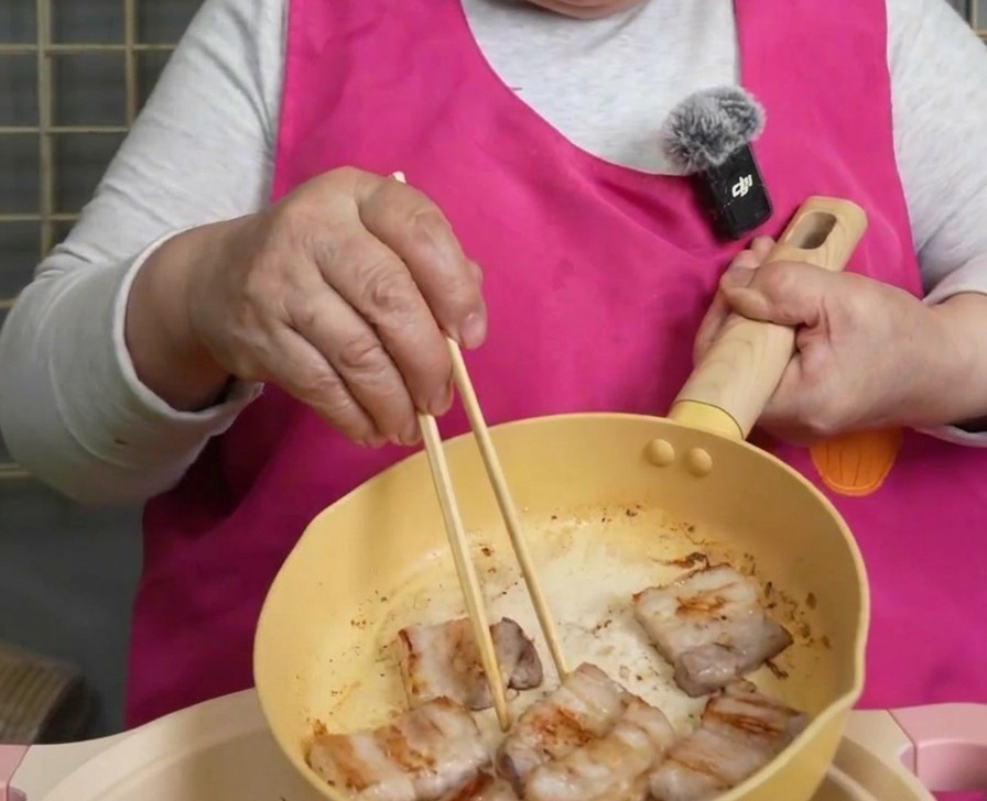 Slices of browned and crispy pork belly being lifted from a yellow frying pan with wooden chopsticks.