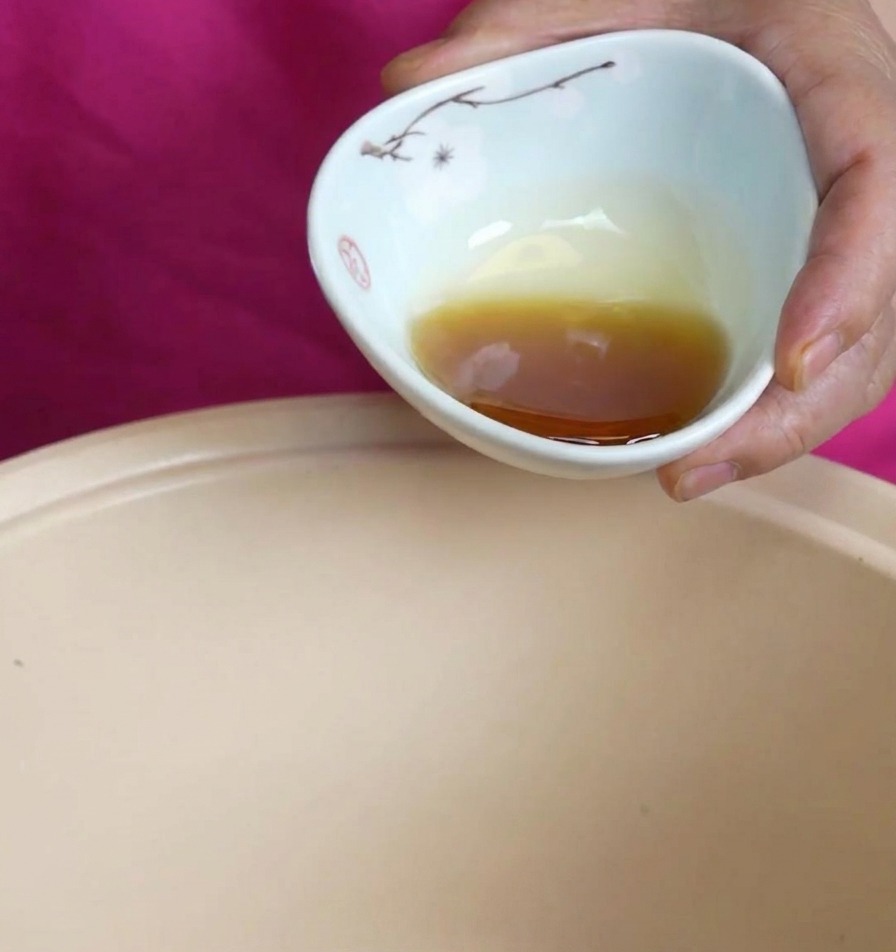Rich, amber-colored sesame oil being poured from a small floral ceramic bowl into a beige cooking pot.