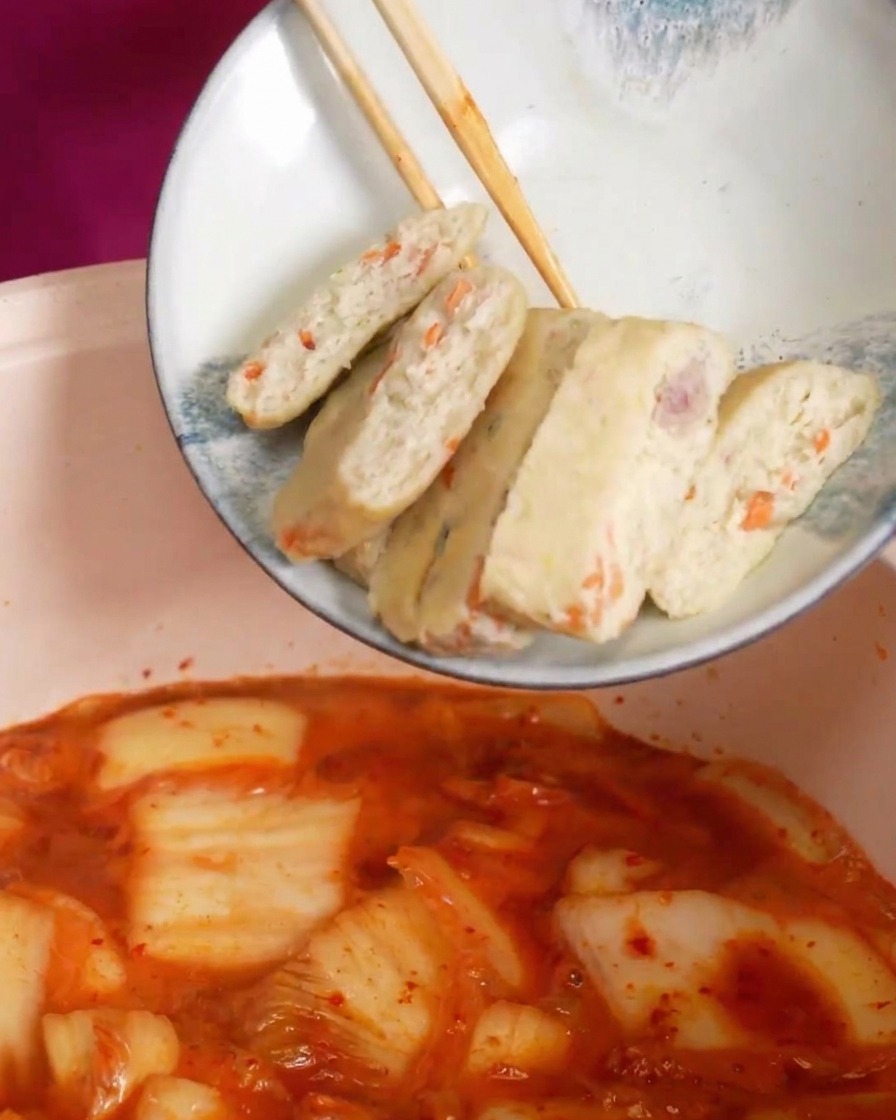 Slices of pale fish cakes with orange vegetable bits being dropped into a bubbling, bright red kimchi soup.