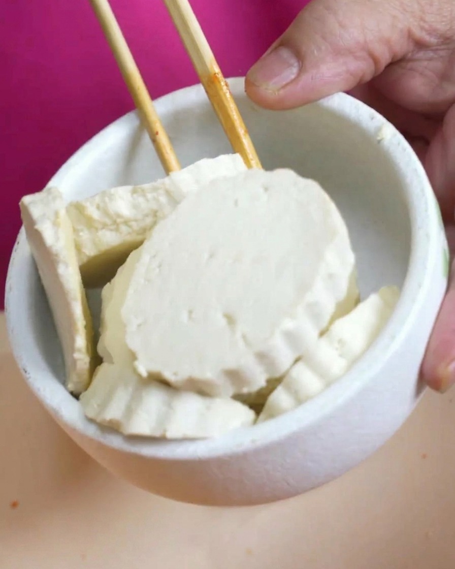 A small white bowl filled with thick, round slices of fresh white tofu being held over a pot of soup.