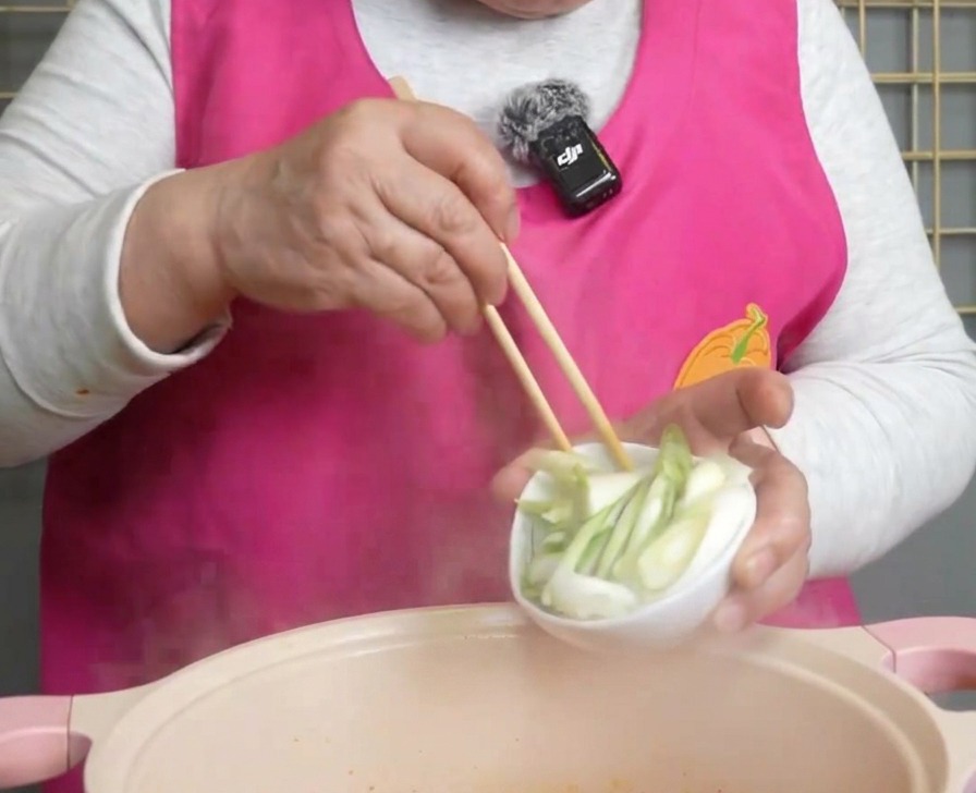 Freshly chopped green onions being poured from a small white bowl into a steaming pot of red kimchi soup.