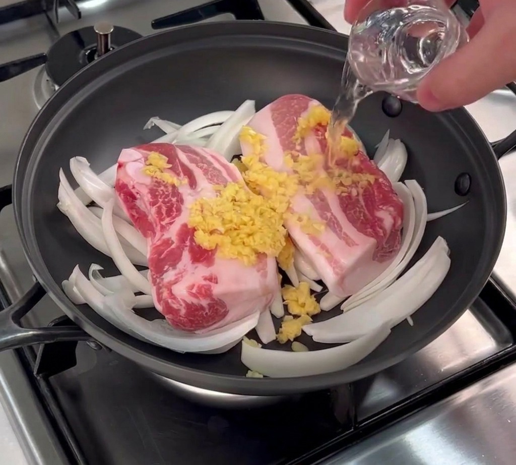A small glass of clear rice wine being poured over raw whole pork belly slabs topped with minced garlic in a black pan.