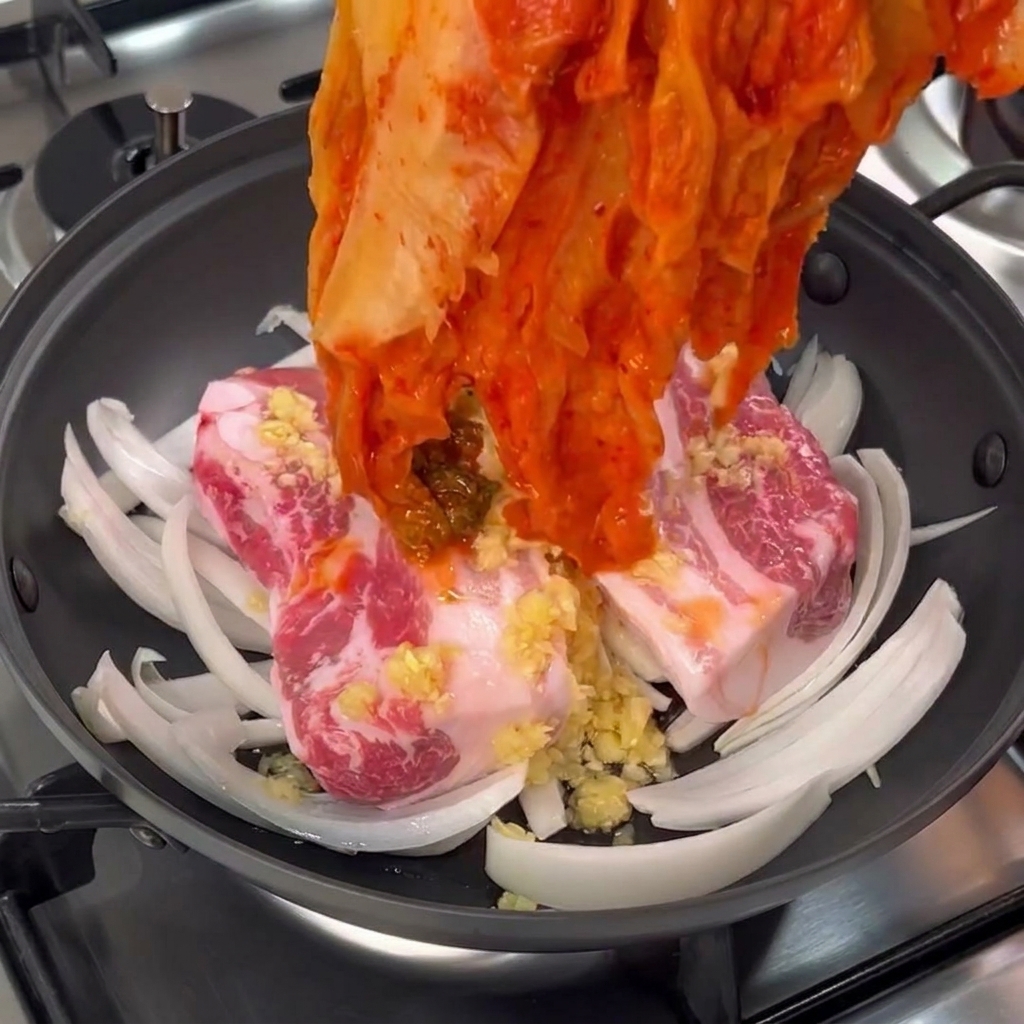 A hand placing a large bundle of vibrant red aged cabbage kimchi over raw pork belly pieces in a pan.