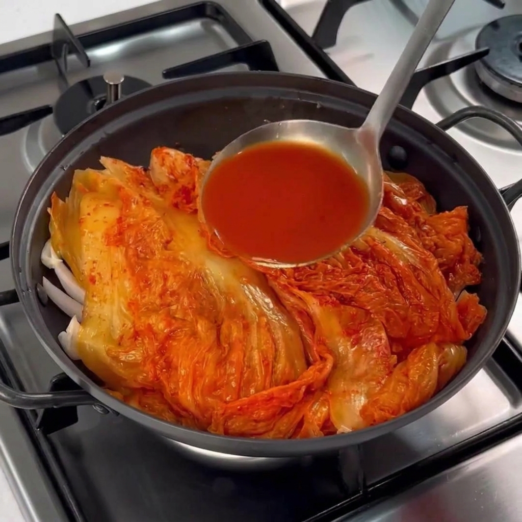 A metal ladle pouring bright red kimchi brine over a pile of aged kimchi sitting on pork belly in a black cooking pot.