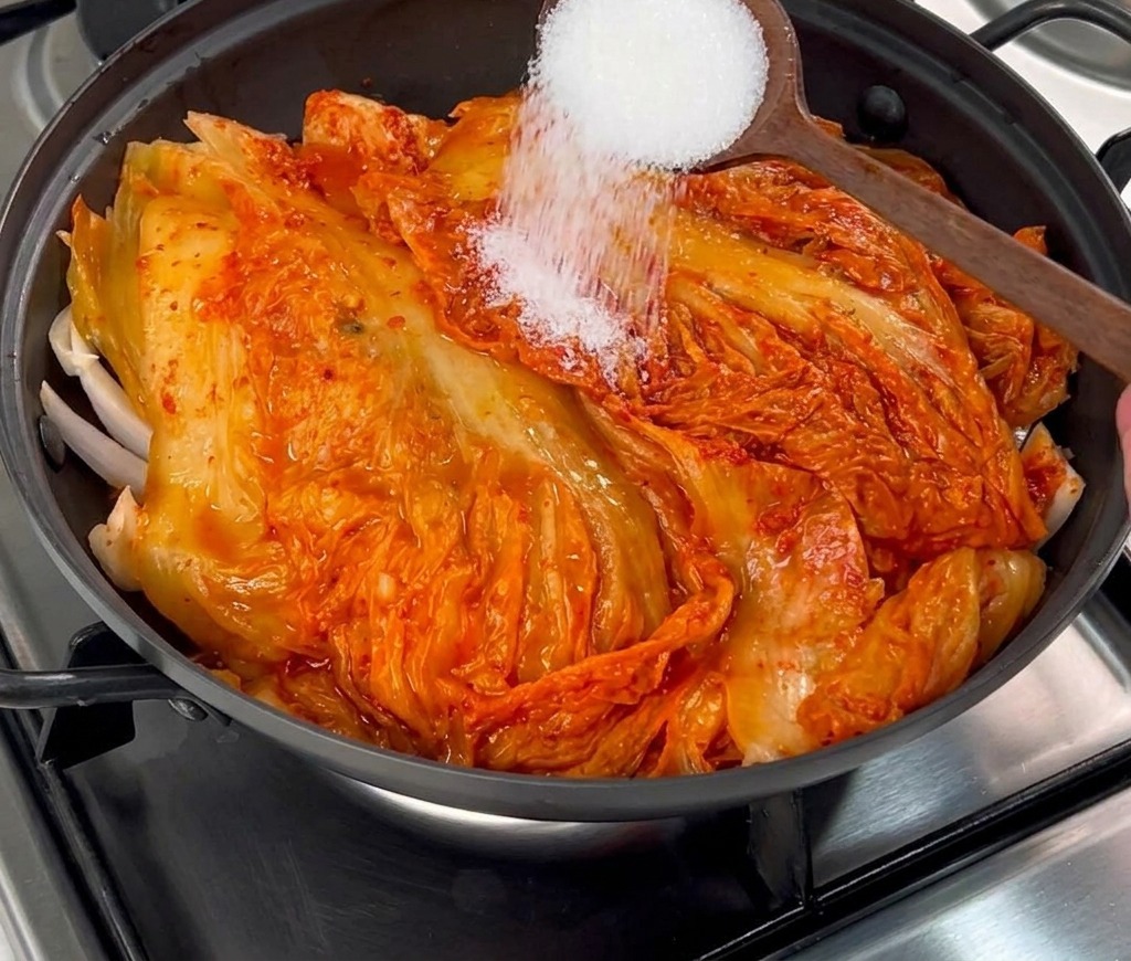 White granulated sugar being sprinkled from a wooden spoon onto red aged kimchi in a cooking pot.