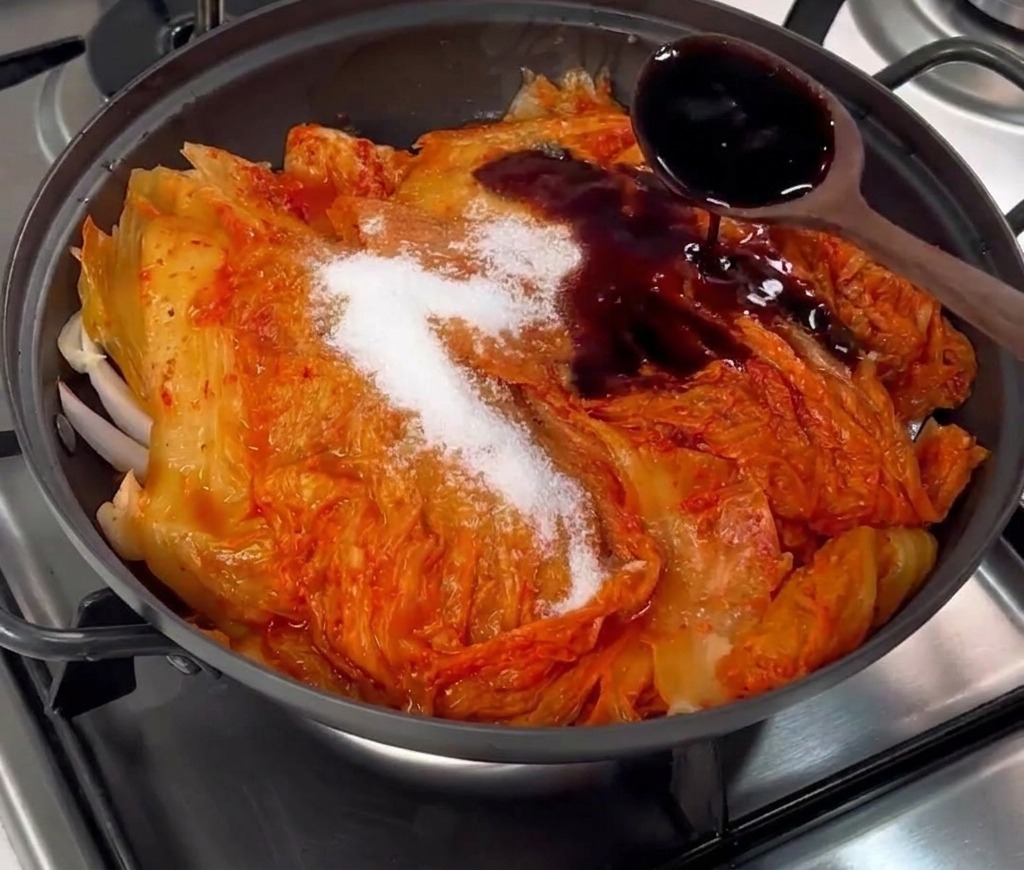 Dark liquid soy sauce being poured from a wooden spoon over kimchi and sugar in a black pan.