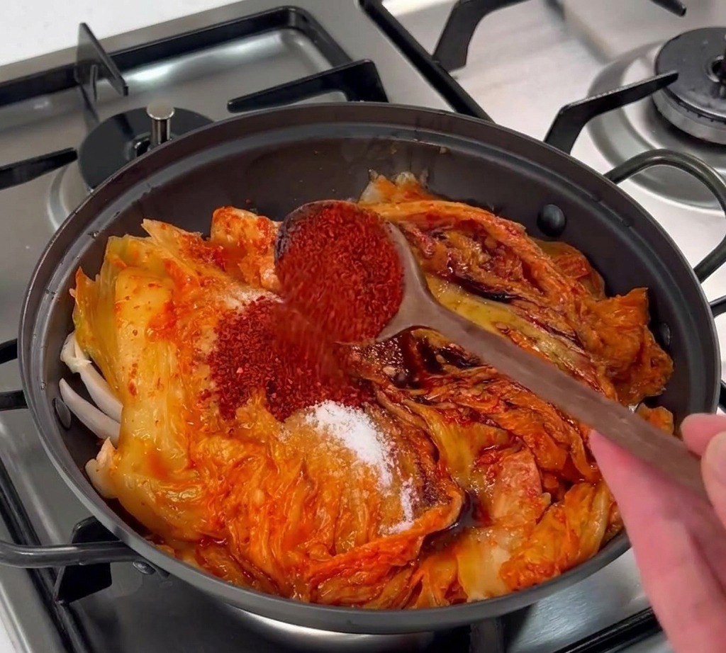 A wooden spoon pouring bright red Korean chili flakes over a layer of fermented kimchi in a pot.