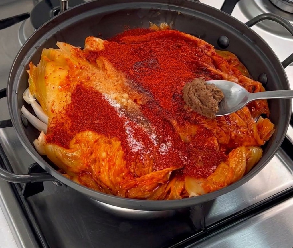 A small dollop of thick brown soybean paste on a metal spoon being placed over a layer of red chili powder and kimchi.