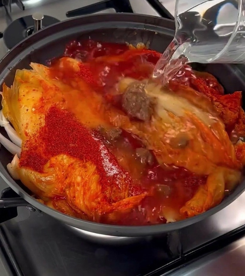 Clear water being poured from a glass pitcher into a pot containing aged kimchi, pork belly, and red chili flakes.