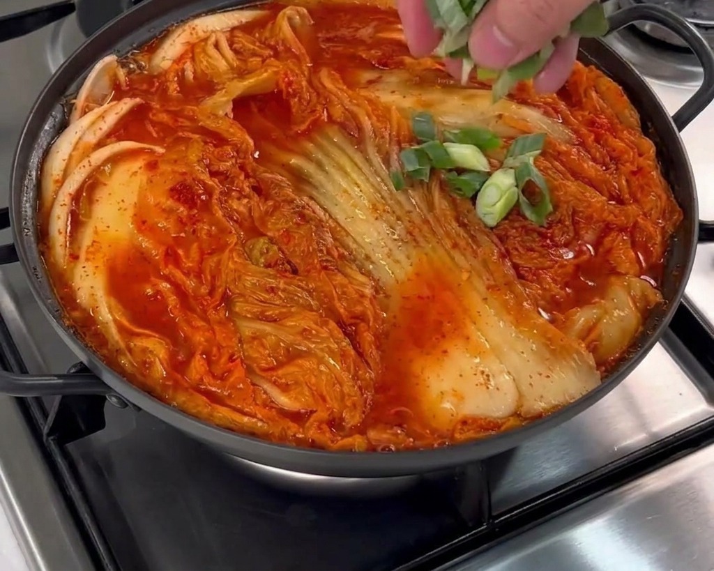 A hand scattering freshly chopped green scallions over a pot filled with kimchi, pork, and red broth.