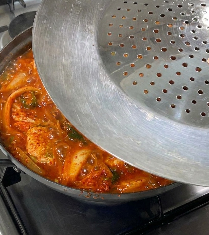 A hand placing a metal lid with small holes over a bubbling pan of kimchi and pork belly stew on a gas stovetop.