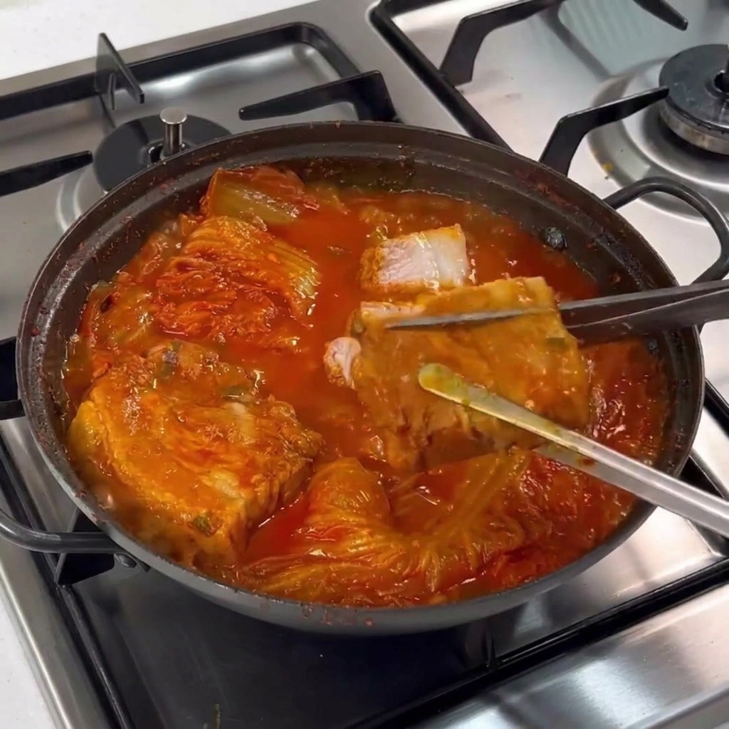Metal tongs holding a piece of cooked pork belly while stainless steel scissors cut it into slices inside a bubbling stew.
