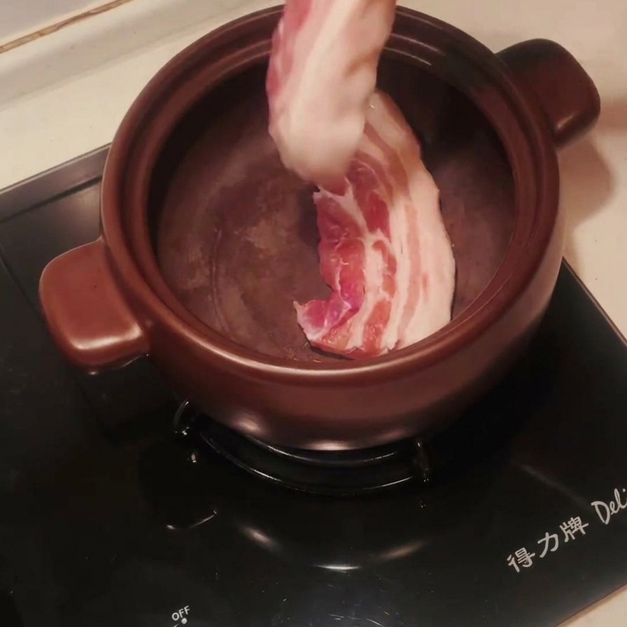 Raw pork belly strips being placed into a preheated dark clay pot with metal tongs.