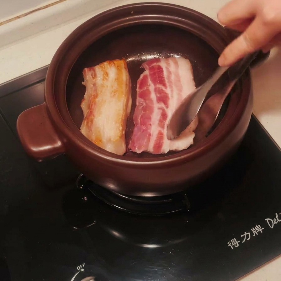 Pork belly strips in a clay pot being flipped with tongs, showing one side perfectly browned and golden.